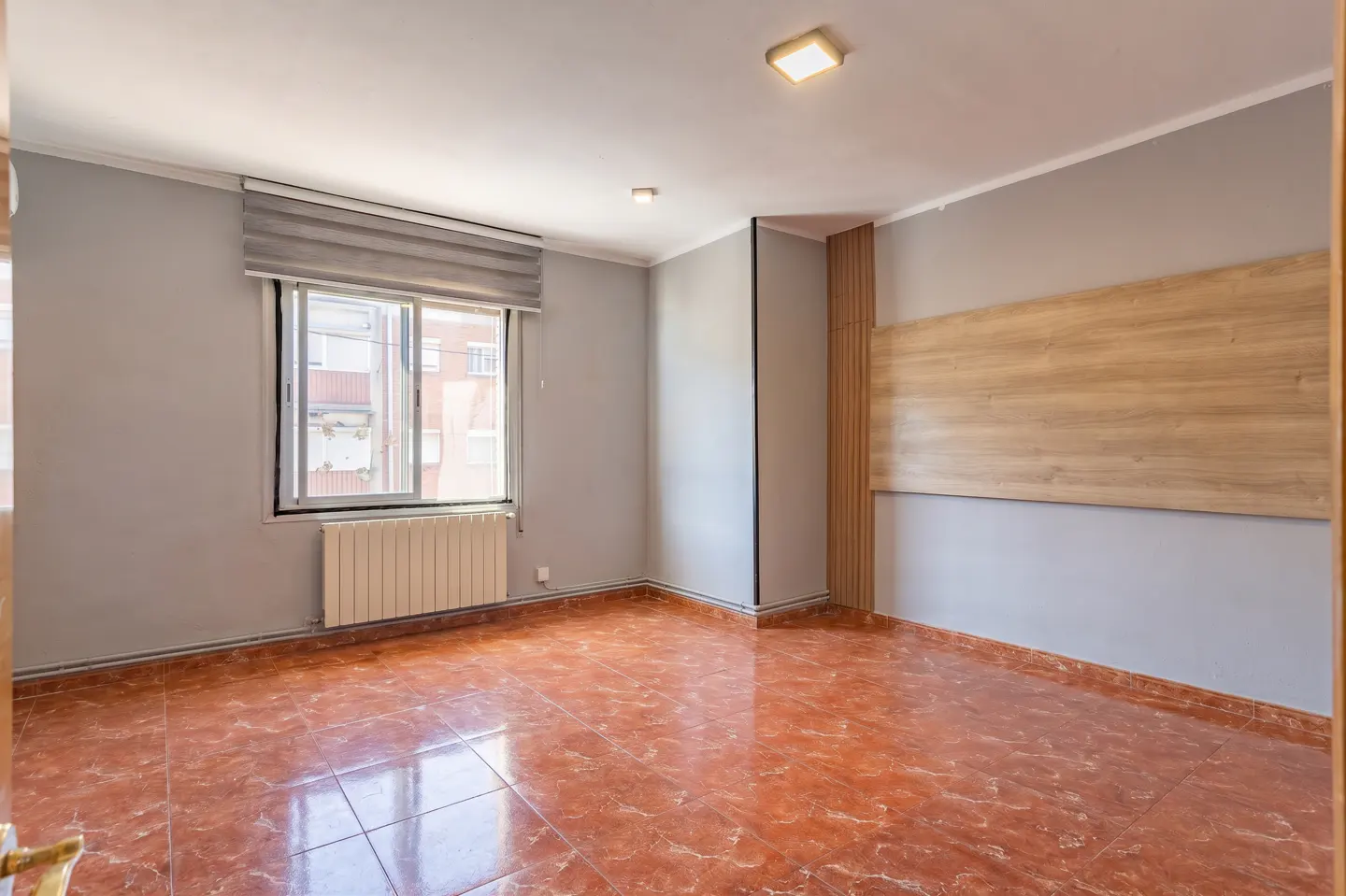 Empty room with grey walls, reddish-brown tiled floor, a window with blinds, a radiator, and a wood-paneled accent wall.