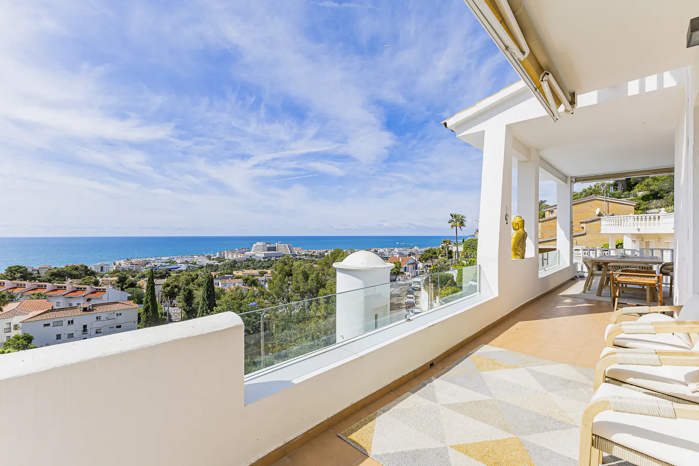 Balcony view of ocean and city. White walls, glass railing, patterned floor. Lounge chairs and dining table visible. Blue sky.