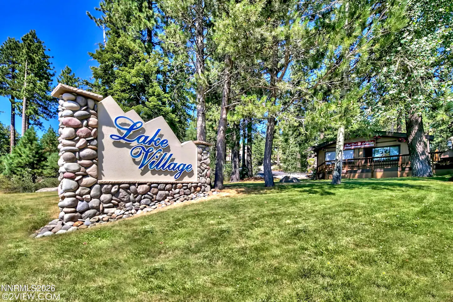 Lake Village sign with blue lettering, stone base, and green lawn. Trees and a building are in the background.