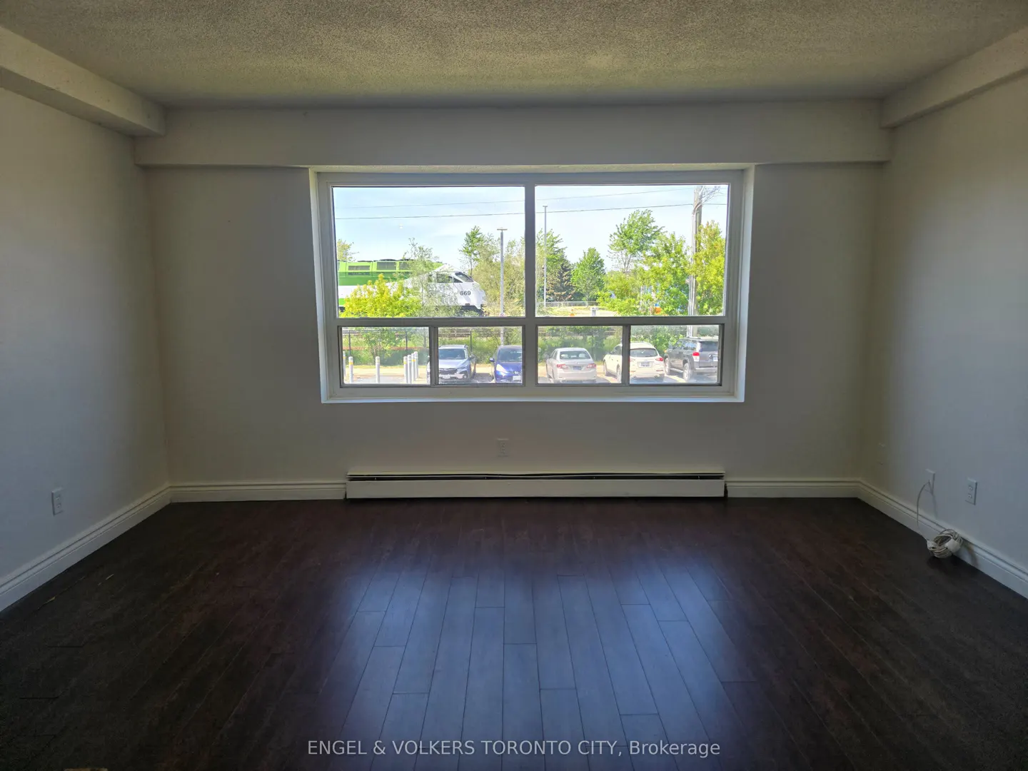 Empty room with dark hardwood floors, white walls, and a large window overlooking a parking lot and a green commuter train.