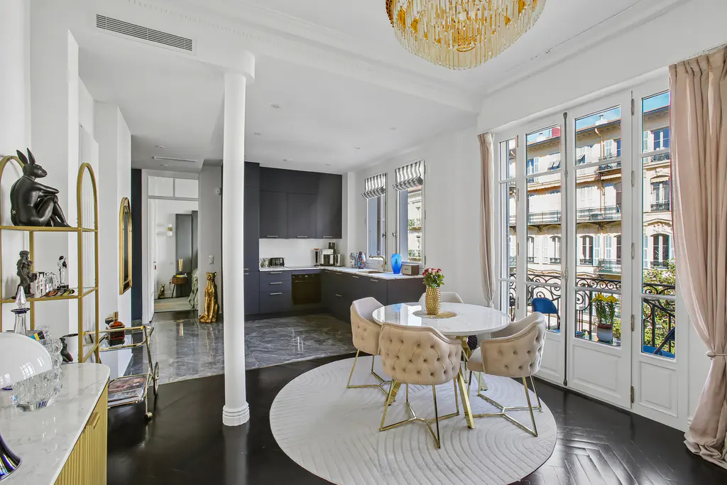 Bright, modern apartment with black floors, white walls, and a gold chandelier. A round table with beige chairs sits on a white rug near French doors.