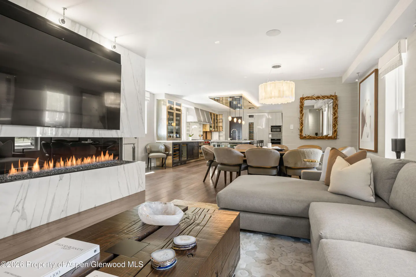 Open-concept living space with a fireplace, TV, and a gray sectional sofa. A dining table and kitchen are in the background.