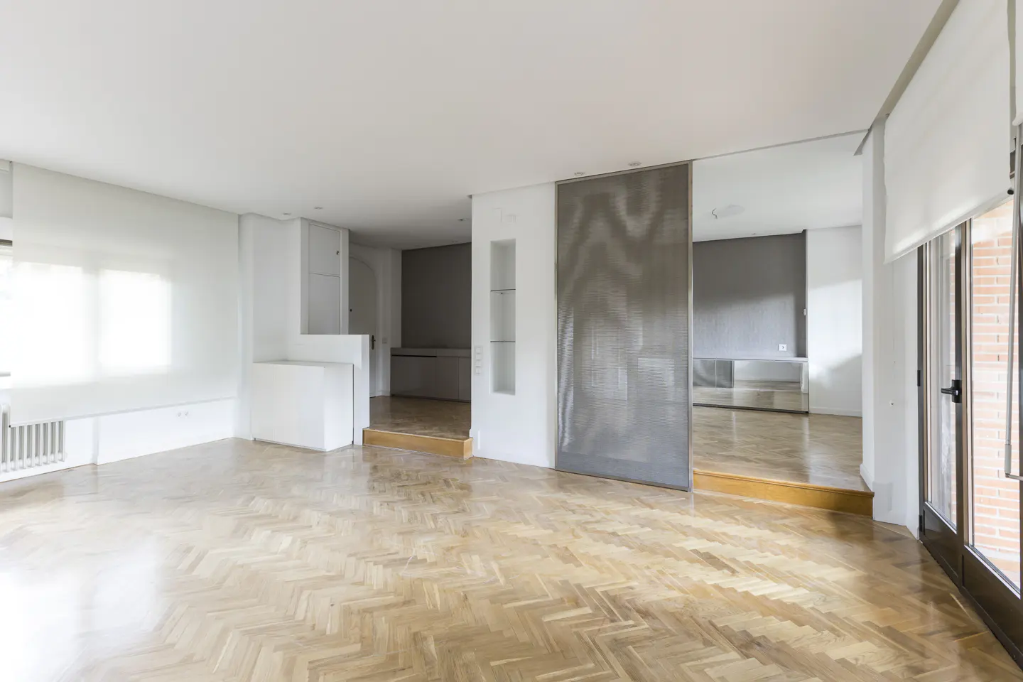 Bright, empty room with herringbone wood floors, white walls, and large windows. A gray metal screen divides the space.