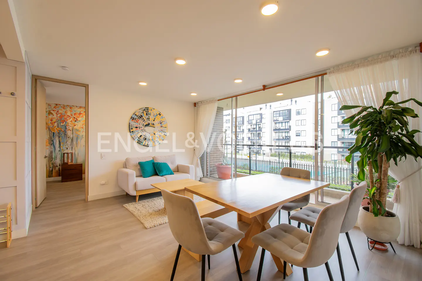 Bright, modern apartment with a dining table, sofa with teal pillows, and a balcony view of a building complex.