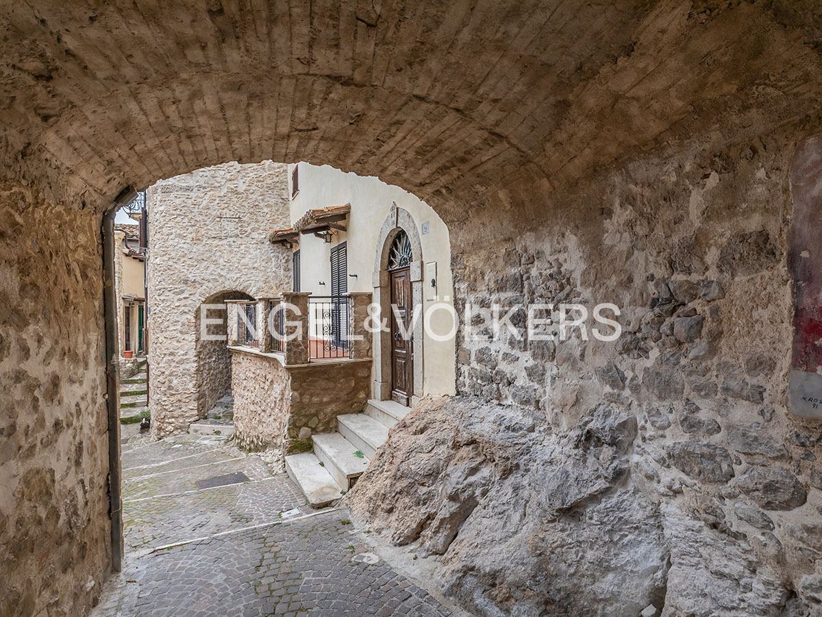 Stone archway frames a narrow street in an old European town. A house with a brown door and stone steps is visible.
