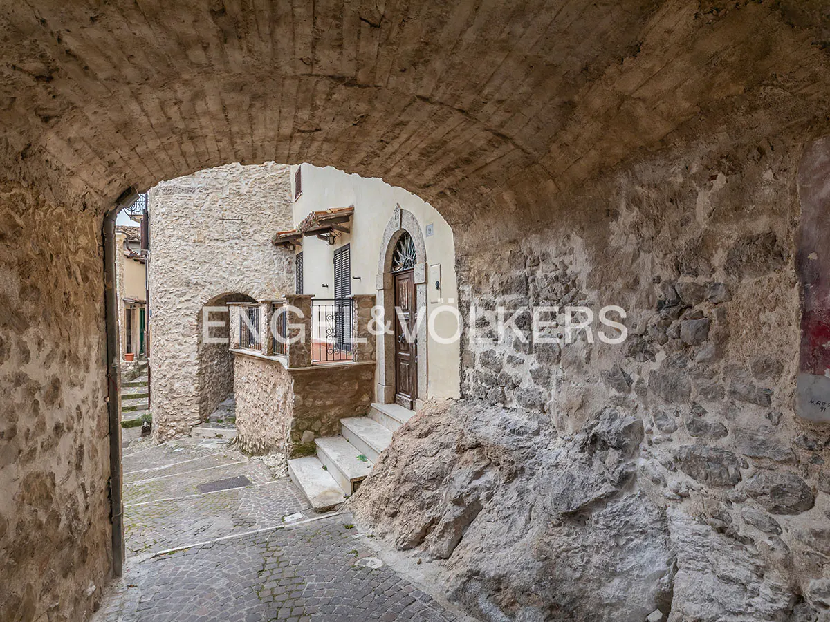 Stone archway frames a narrow street in an old European town. A house with a brown door and stone steps is visible.