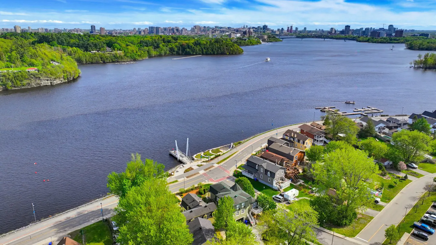 Aerial view of houses along a riverbank with a boat dock, lush green trees, and a distant city skyline under a blue sky.