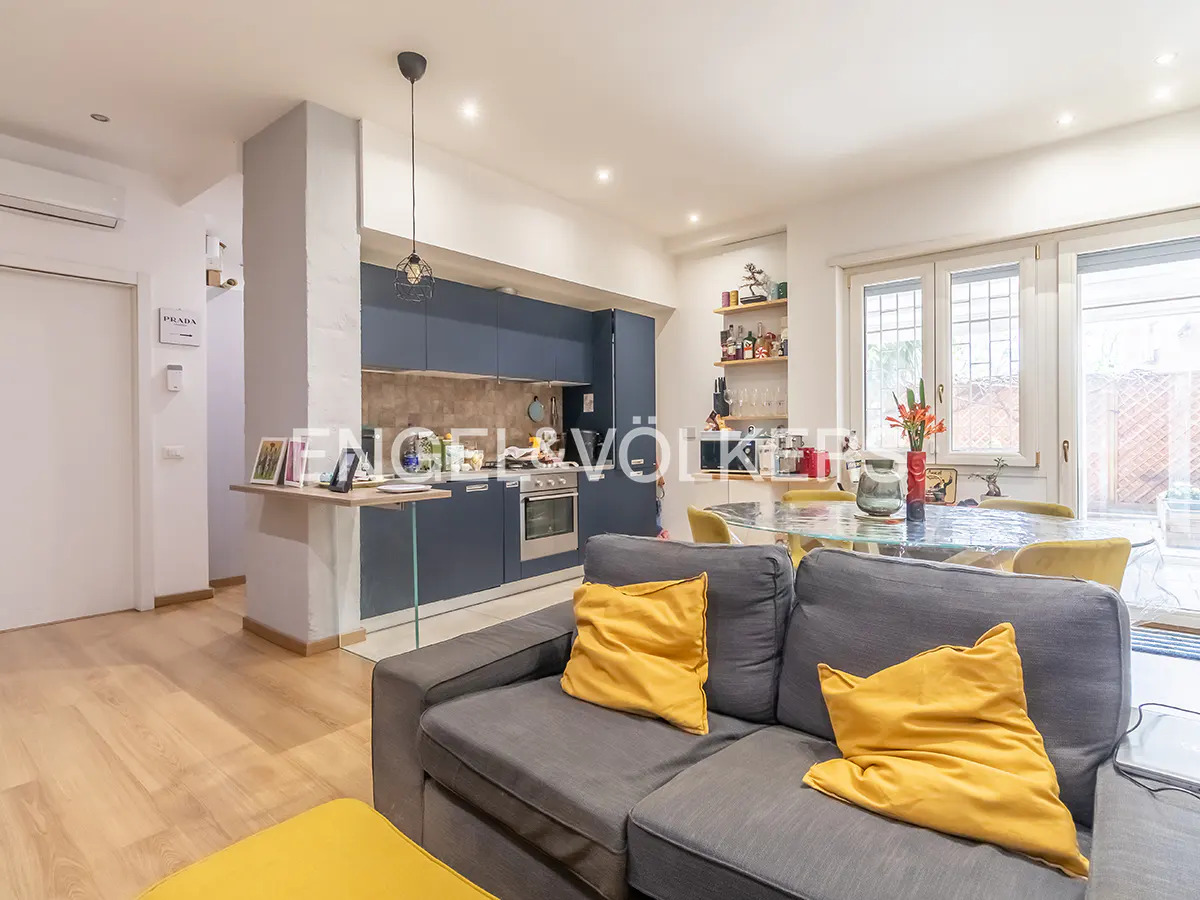 Open-concept living space with a gray sofa, yellow pillows, and a blue kitchen with stainless steel appliances. A glass table and chairs are in the background.