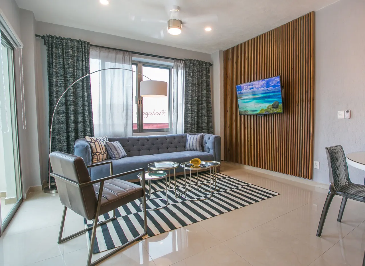 Living room with a blue sofa, striped rug, and wooden accent wall with a TV. A modern chair and round tables complete the space.