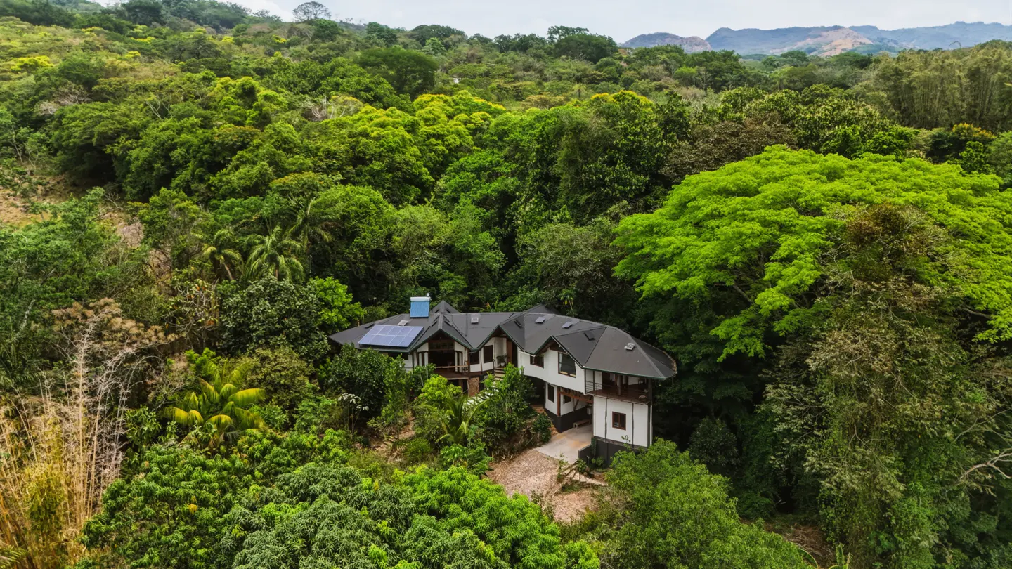 Aerial view of a modern white house with a black roof, nestled in a lush green forest with mountains in the distance.