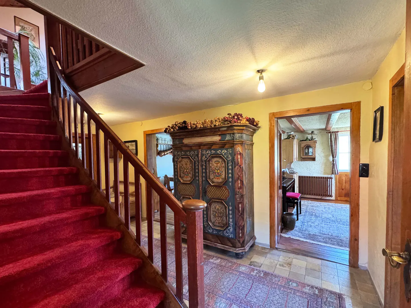 Interior view of a home's foyer with red carpeted stairs, a painted armoire, and a view into another room.
