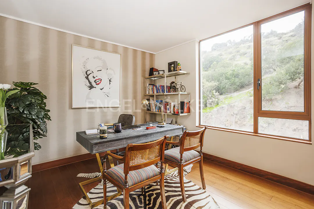 A home office with a desk, two chairs, and a large window with a view of a green hillside. A zebra-print rug is on the wood floor.