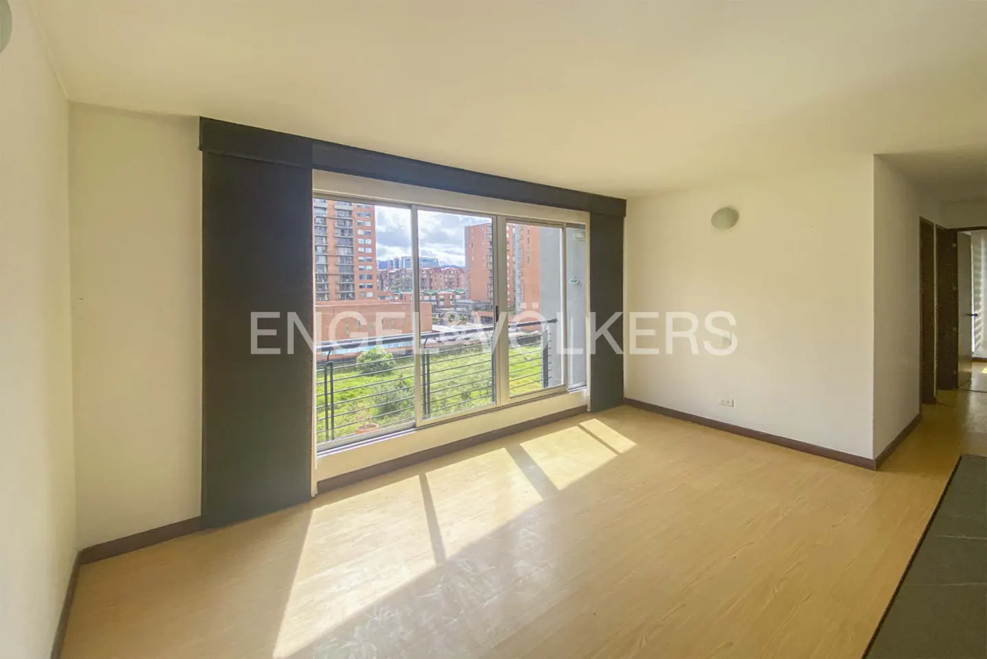 Empty room with light wood floors, cream walls, and a large window overlooking a city view with brick buildings.