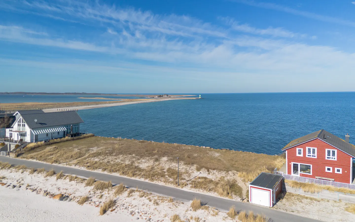 Aerial view of a white and red house near a blue sea with a lighthouse in the distance under a blue sky.