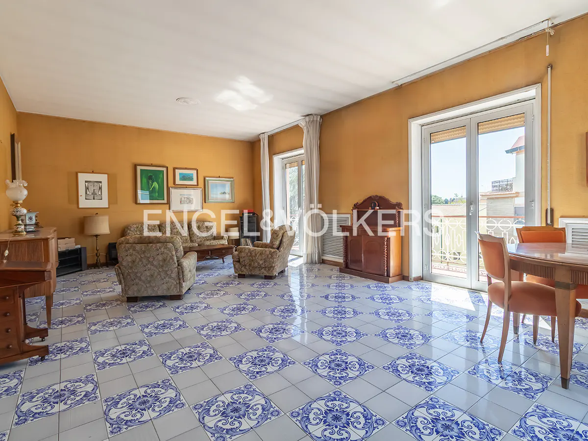 A spacious living room with yellow walls, blue and white tiled floors, and a balcony.
