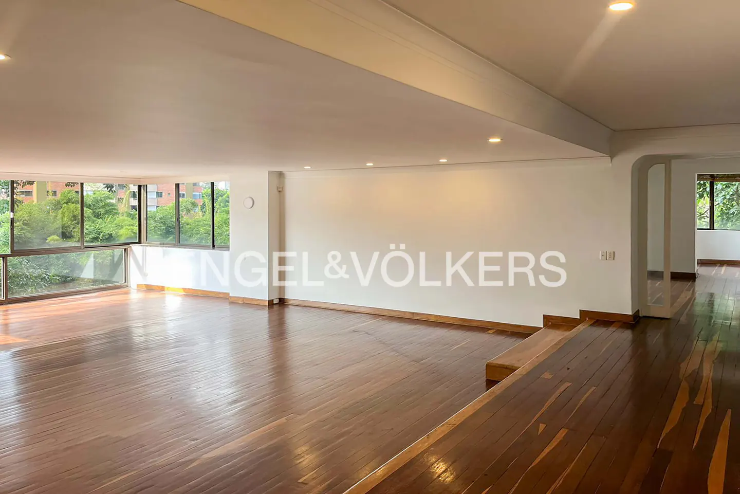 Bright, empty living room with hardwood floors, white walls, and large windows overlooking green trees. Engel & Völkers logo on the wall.