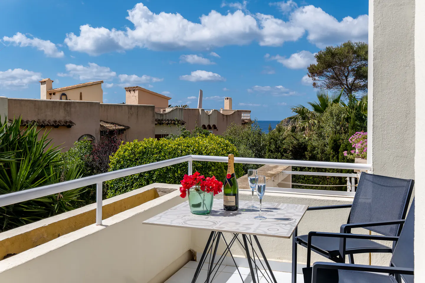 Balcony view with champagne, red flowers, and two chairs. Buildings and blue sky with clouds in the background.
