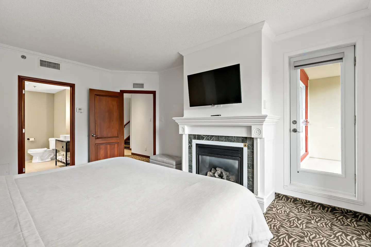 Bedroom with white bedding, fireplace, TV, and balcony door. Bathroom and stairs are visible through open doors.