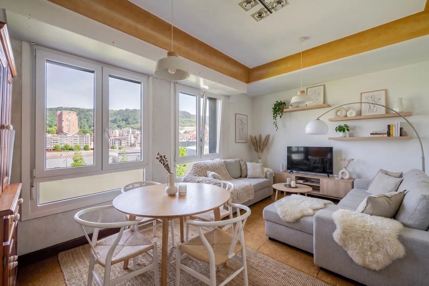 Bright living room with a round wooden table, white chairs, and a gray sofa. Large windows offer a city view. Shelves and decor add a cozy touch.