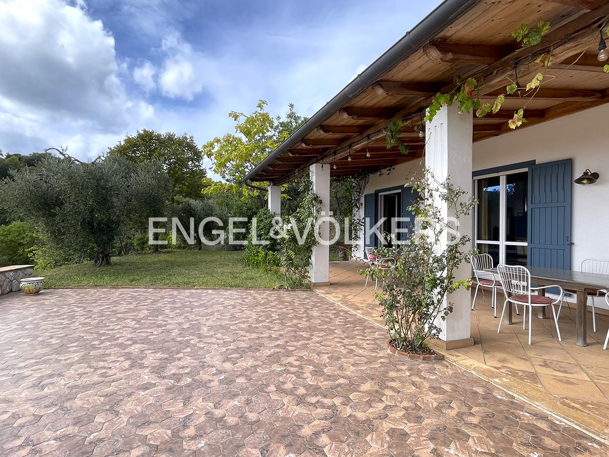 Spacious stone-paved patio with outdoor dining set under a wooden pergola, overlooking a lush garden with olive trees. [AI-generated text]