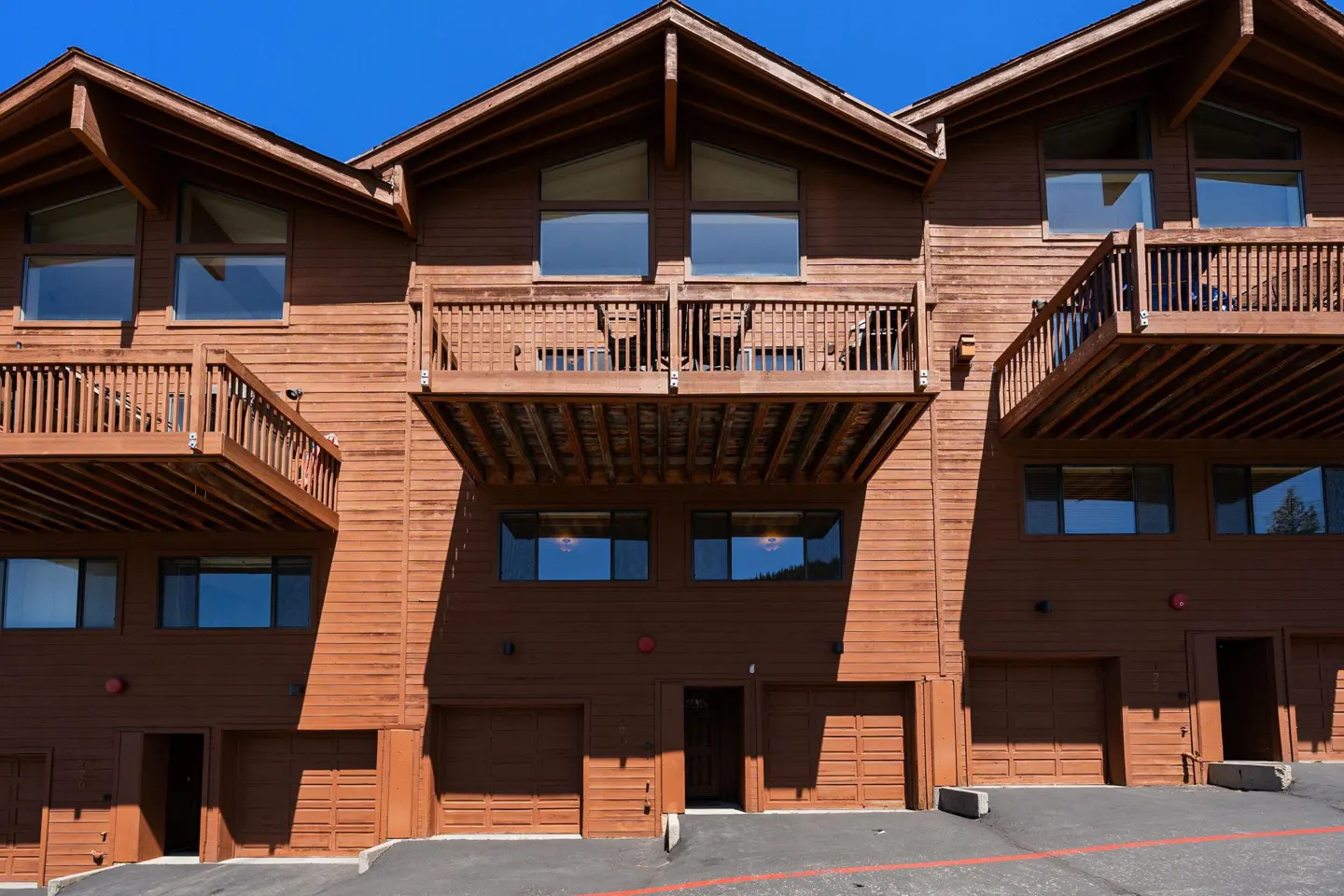 Exterior view of a multi-story brown wooden townhouse complex with balconies and ground-level garages under a clear blue sky.