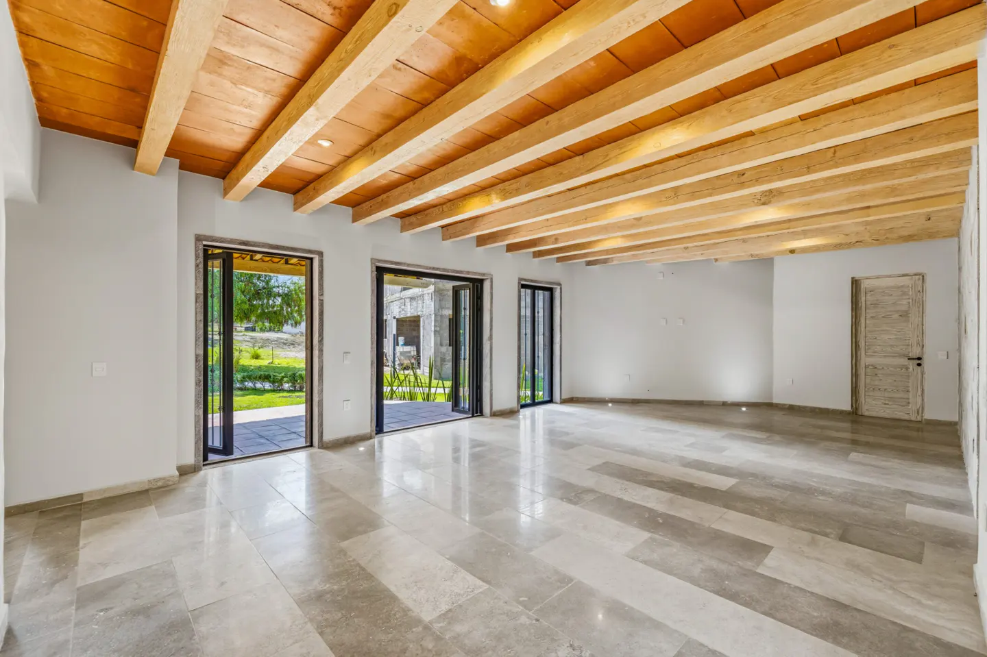 Bright, empty room with gray tile floor, white walls, and wood-beamed ceiling. Open doors lead to a green outdoor space.