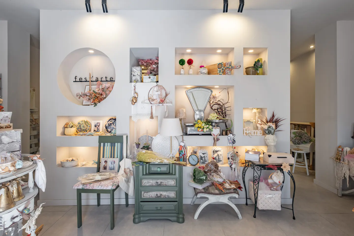 Interior of a shop with white walls and recessed display niches filled with decorative items, furniture, and the "fes'arte" logo.