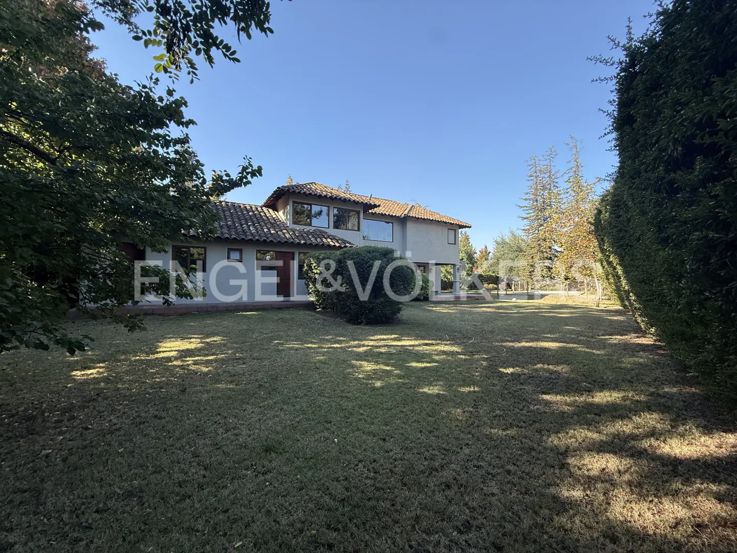 Two-story house with a red tile roof and large lawn, framed by trees and shrubs under a blue sky.