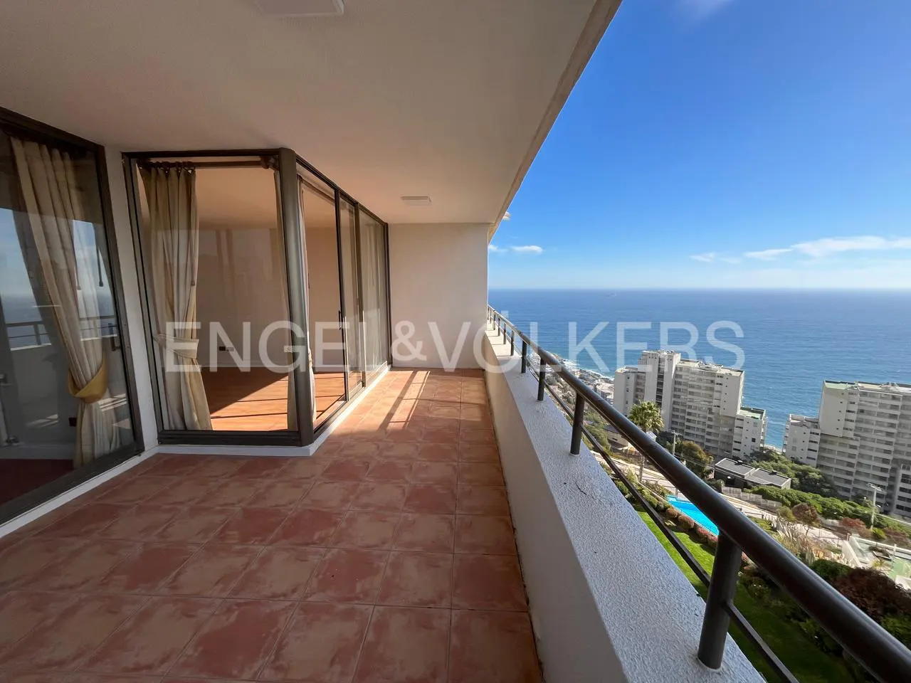 Balcony view of ocean and city. Terracotta tile floor, glass doors with curtains, white walls, and metal railing. Blue sky.