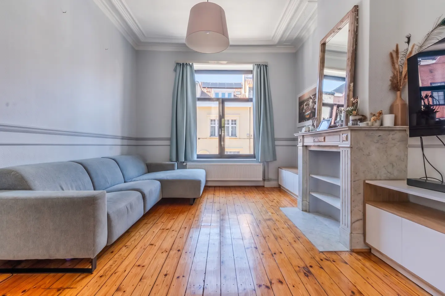 Living room with wood floors, a gray sofa, and a marble fireplace. A window with blue curtains looks out onto a building.