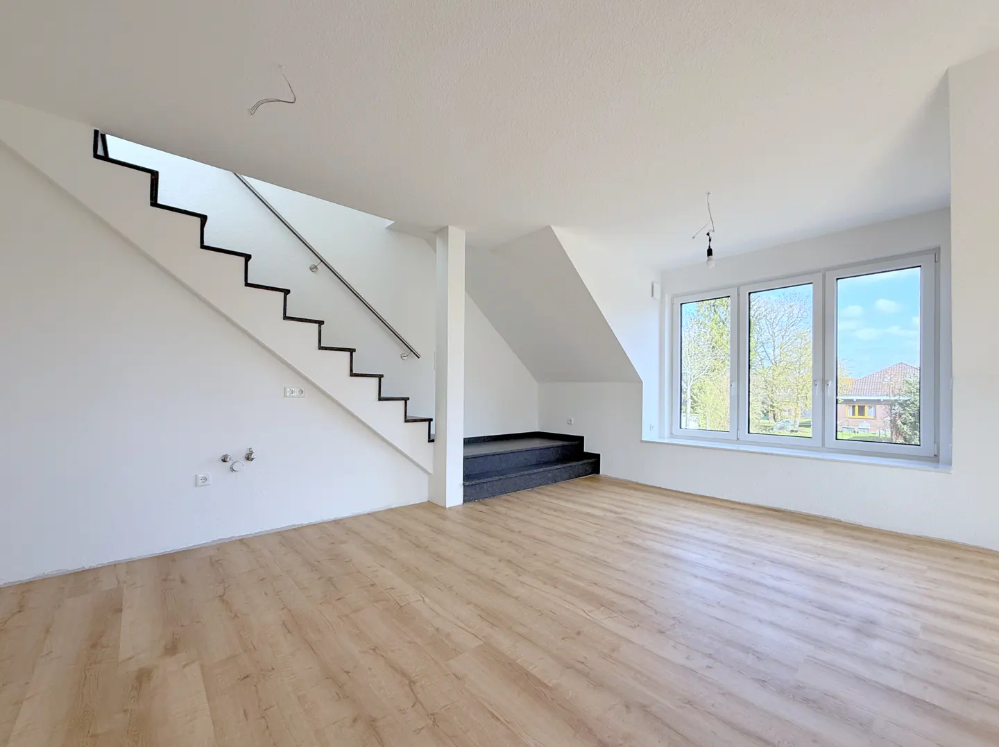 Bright, empty room with light wood floors, white walls, stairs with black trim, and a large window showing trees and a house.