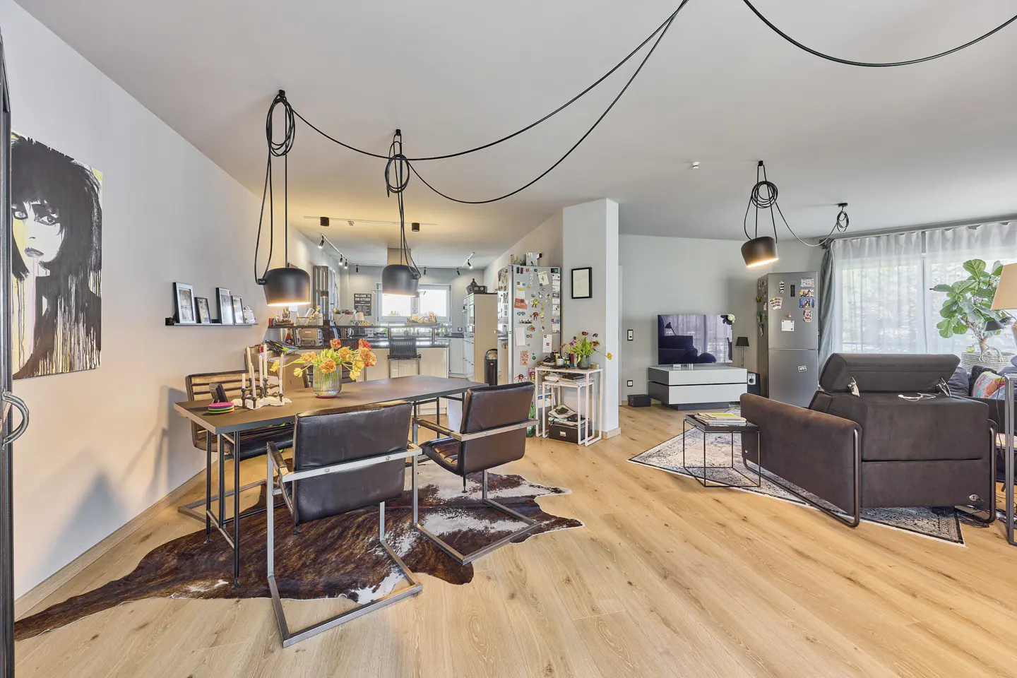 Open-concept living space with wood floors, dining table, and brown leather chairs. A cowhide rug sits under the table. Modern black pendant lights hang above.
