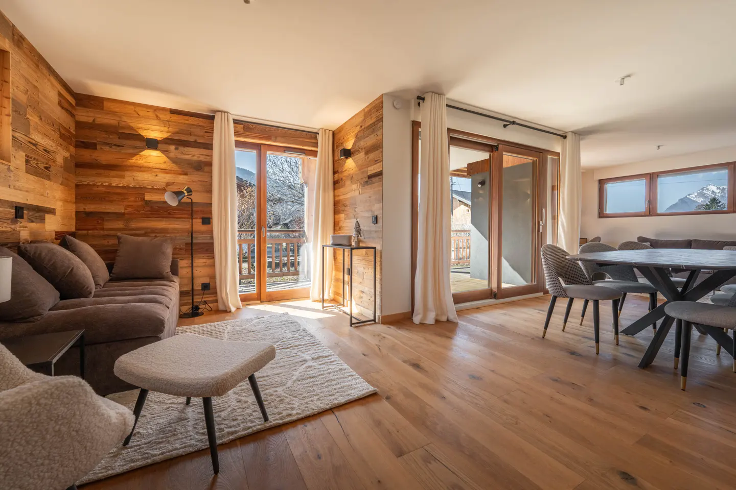 Living room with wood paneled wall, brown sofa, and dining table with chairs, hardwood floors, and balcony doors.