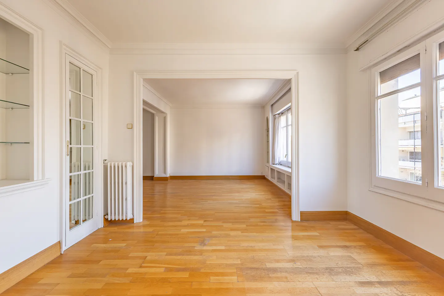 Bright, empty room with hardwood floors, white walls, and trim. A doorway leads to another room, and windows offer a view of a building.