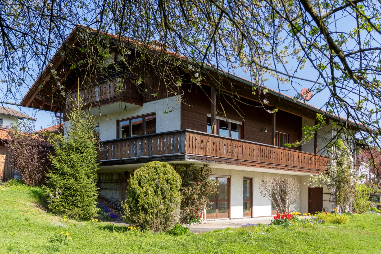 Two-story house with brown trim and a balcony, surrounded by green grass and trees.