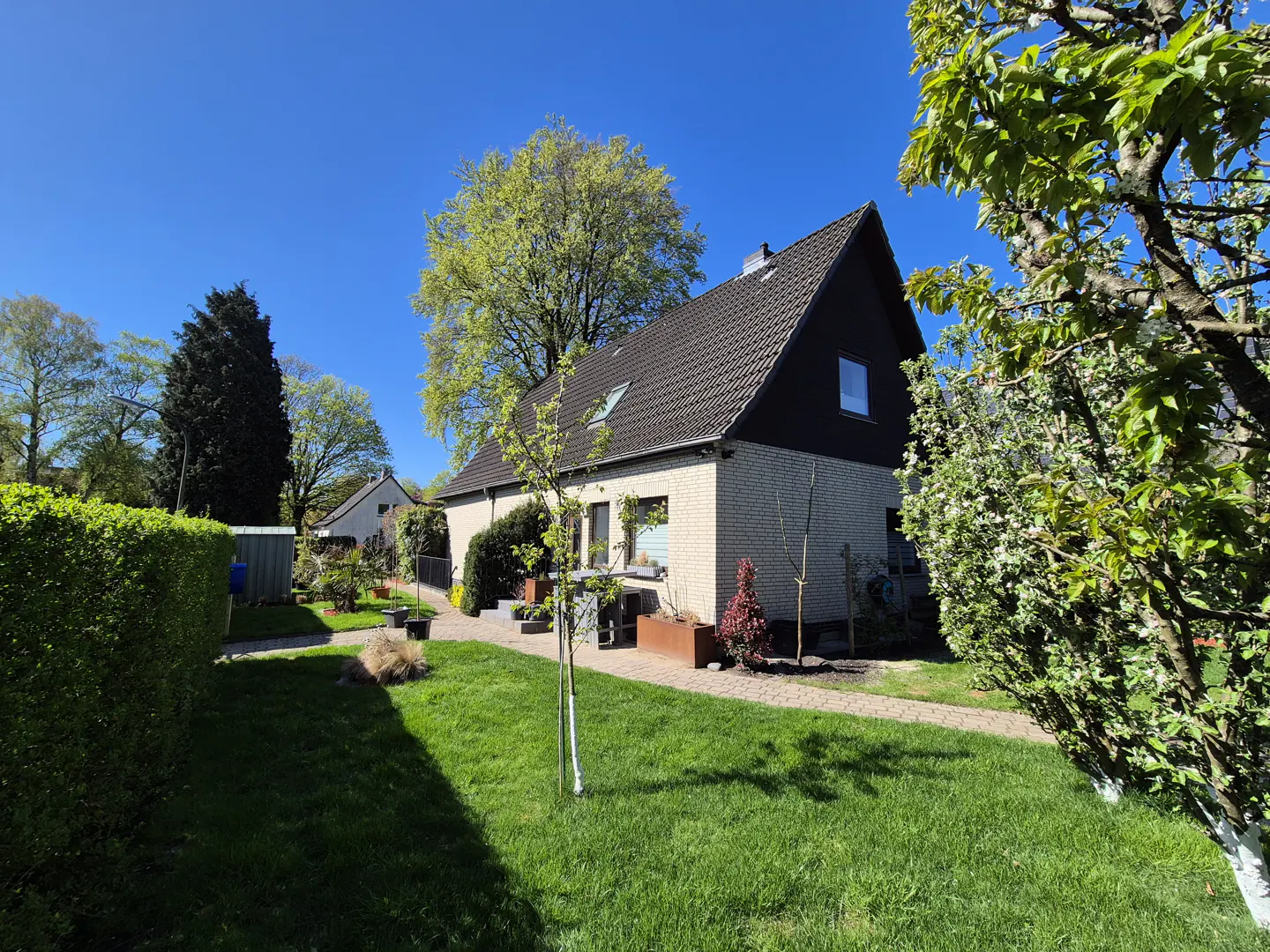 Exterior view of a one-story house with a dark roof, light brick, green lawn, and trees under a blue sky.