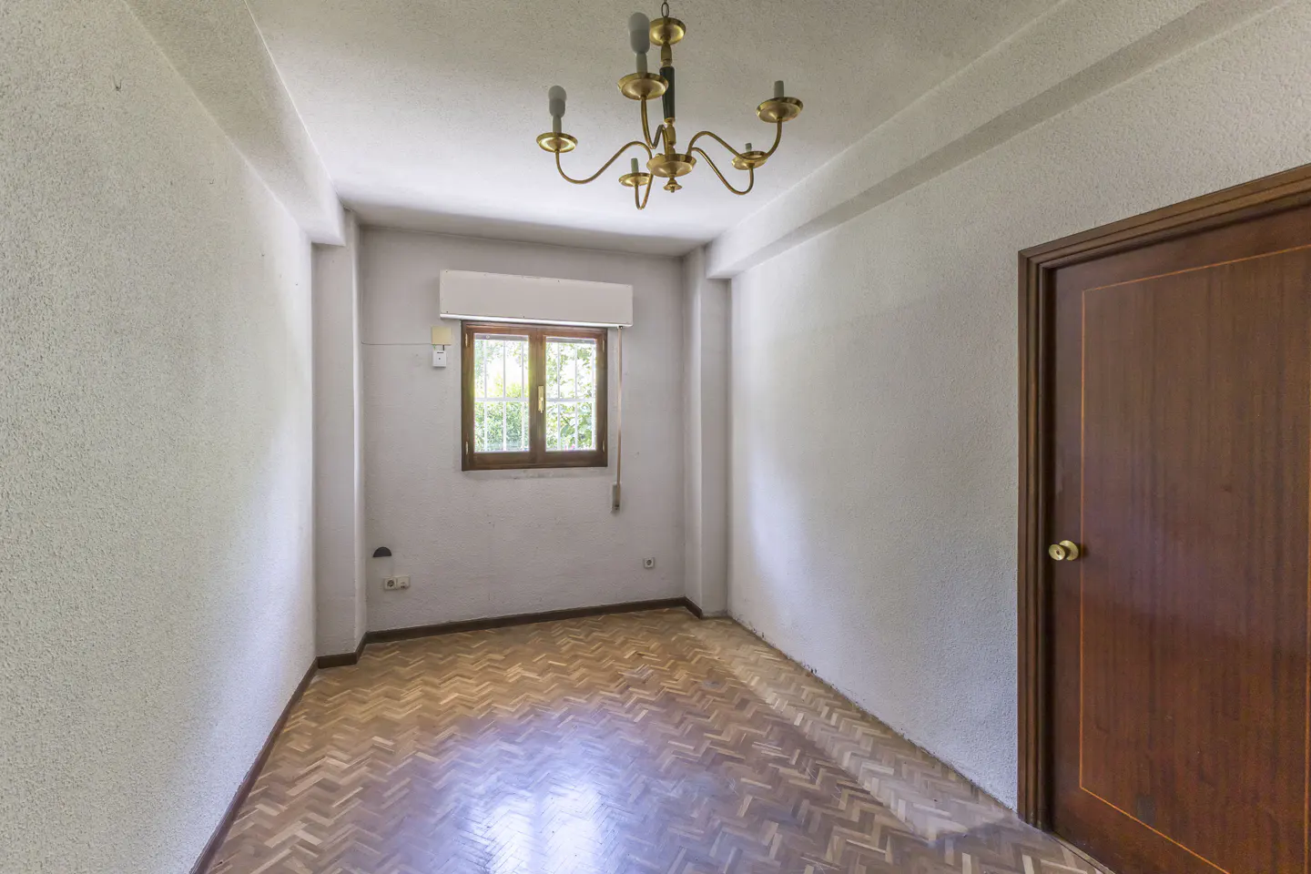 Empty room with white walls, herringbone wood floors, a brass chandelier, and a wooden door on the right.