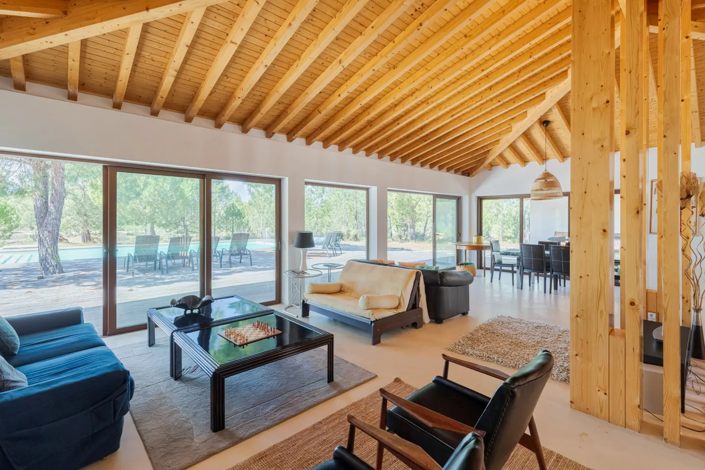 Bright living room with a wooden ceiling, large windows, and a view of the pool. Sofas, chairs, and a chess board are visible.