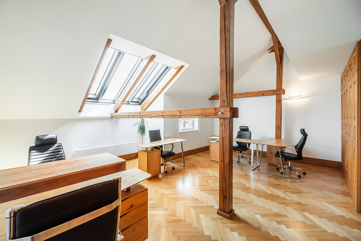 Bright attic office with herringbone wood floors, exposed wooden beams, skylights, and multiple desks with black chairs. [AI-generated text]