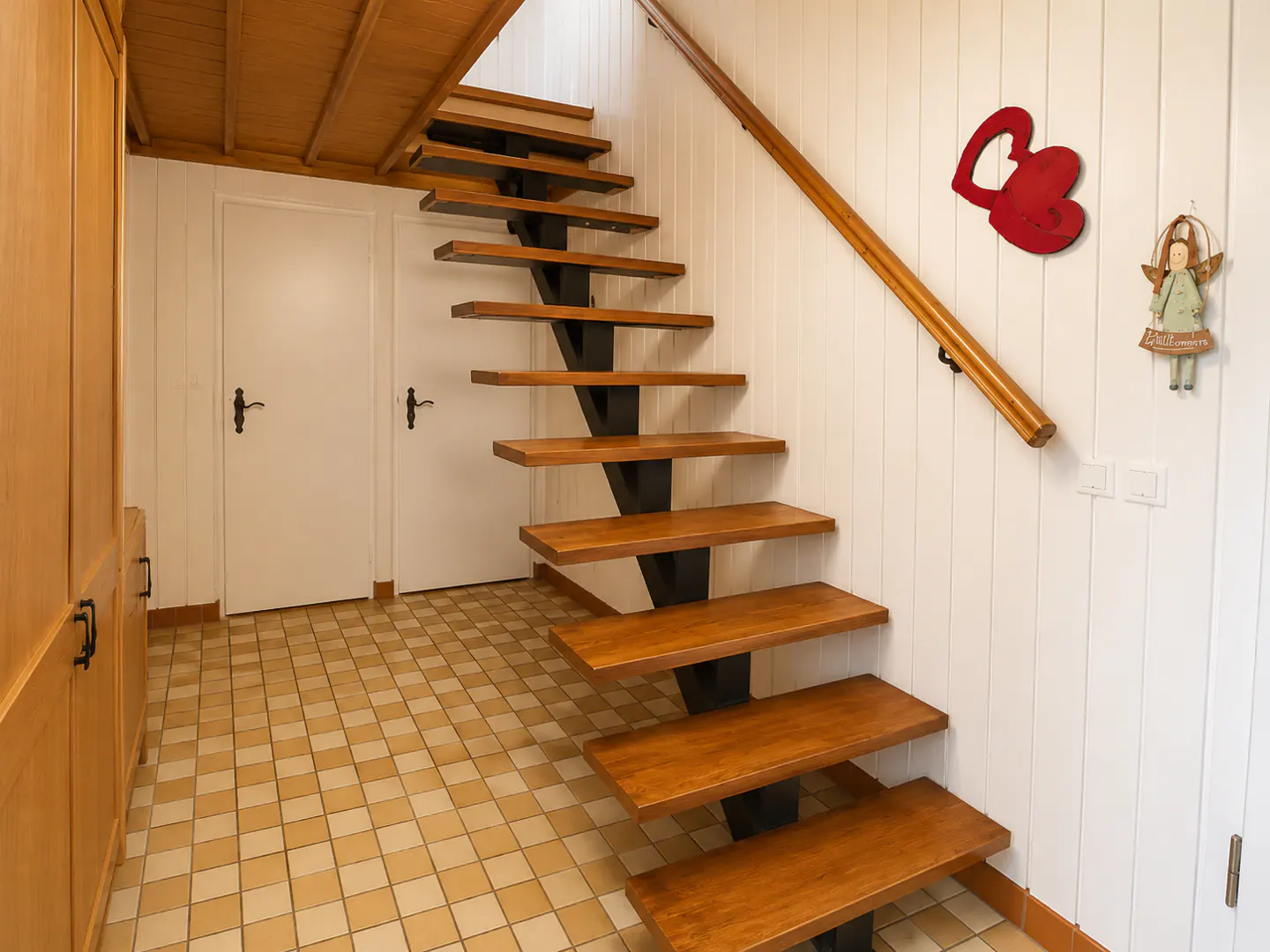 Modern floating staircase with wooden treads and black metal frame in a hallway with tiled floors and white paneled walls.