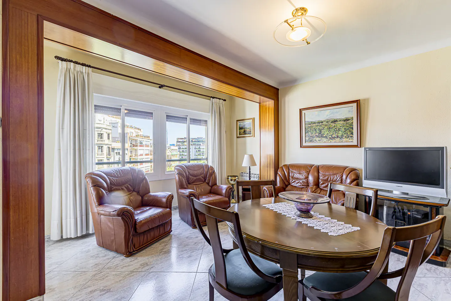 Living room with brown leather armchairs and sofa, a wooden dining table with chairs, and a TV. A window shows a city view.