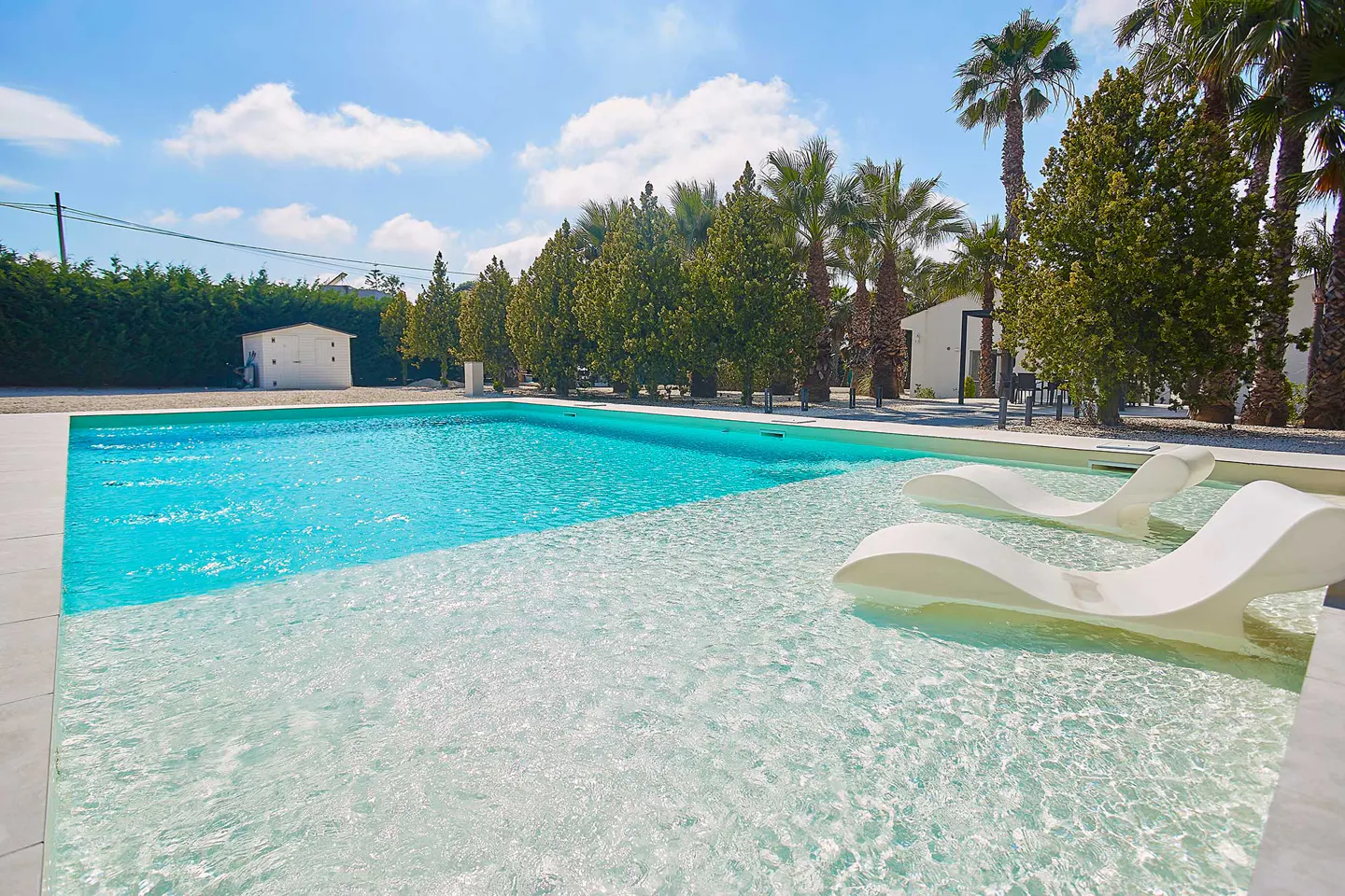 Bright blue swimming pool with a shallow tanning ledge and two white lounge chairs, surrounded by palm trees and greenery.