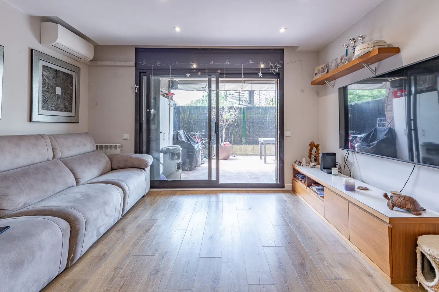 Living room with a gray sofa, wood floors, and sliding glass doors to a patio. A large TV hangs above a long cabinet.