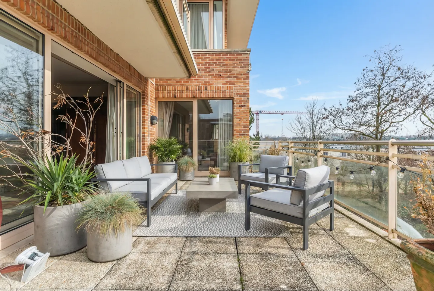 A balcony with gray outdoor furniture, potted plants, and a view of trees and a construction crane under a blue sky.