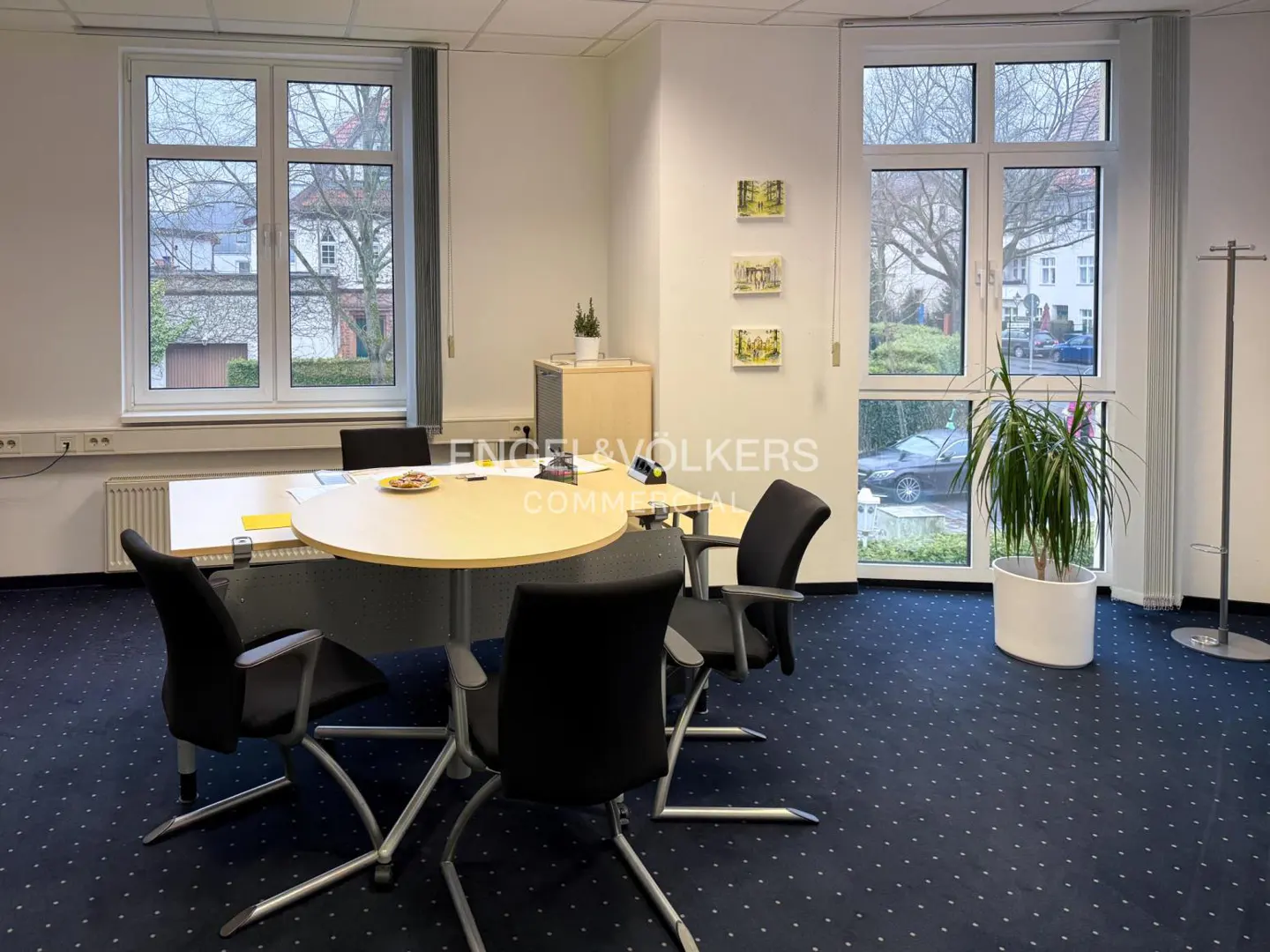 Office space with a round table, black chairs, and large windows. A plant sits near the window, and artwork hangs on the wall. Blue carpet covers the floor.