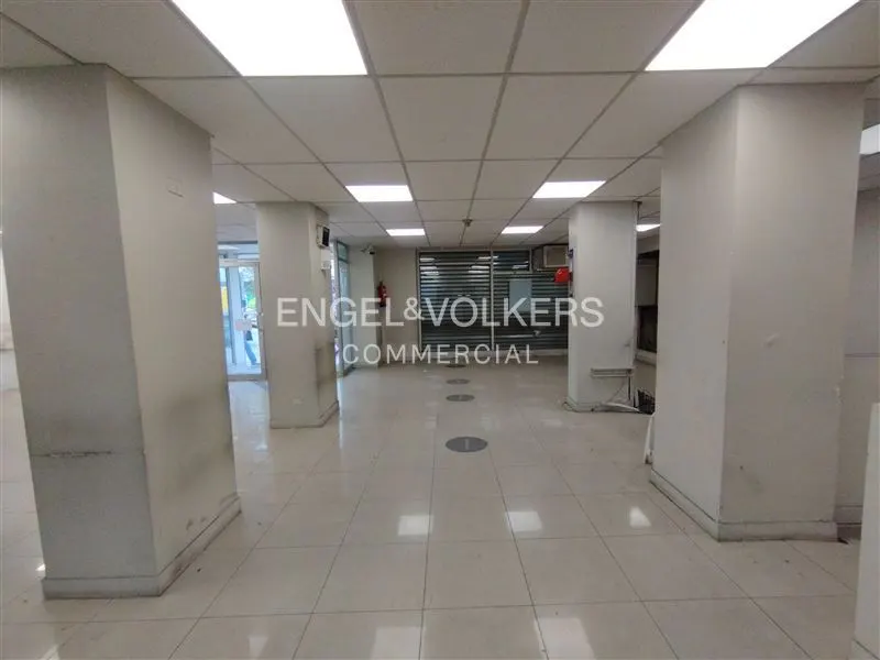 Empty commercial space with white tile floor, white columns, and a white grid ceiling with fluorescent lights. Engel & Volkers logo is visible.