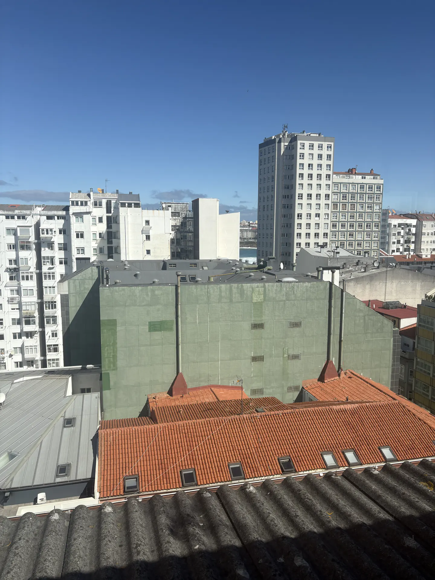 Cityscape view with white apartment buildings, a large green building, and red-tiled roofs under a clear blue sky.