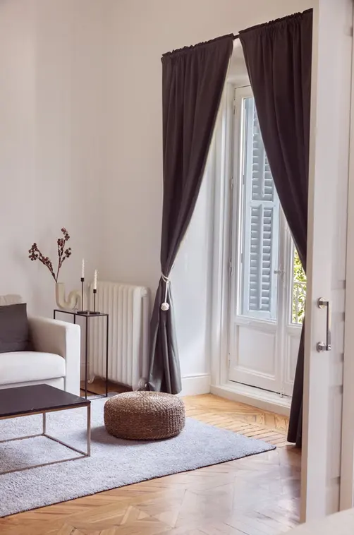 Bright living room with white walls, wood floors, and a gray rug. A white sofa, coffee table, and brown pouf are visible. Dark gray curtains frame a window with white shutters.