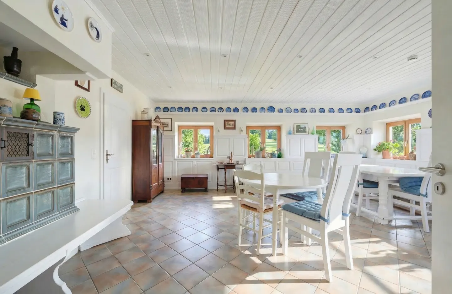 Bright, spacious kitchen with white wood-paneled ceiling, tiled floor, and white furniture. Blue and white decorative plates line the walls.