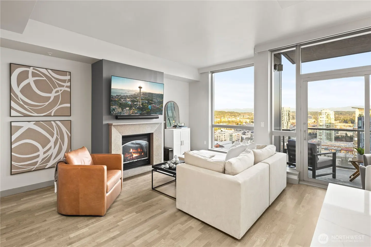 Bright living room with a tan leather chair, white sofa, fireplace with TV, and a city view from large windows.